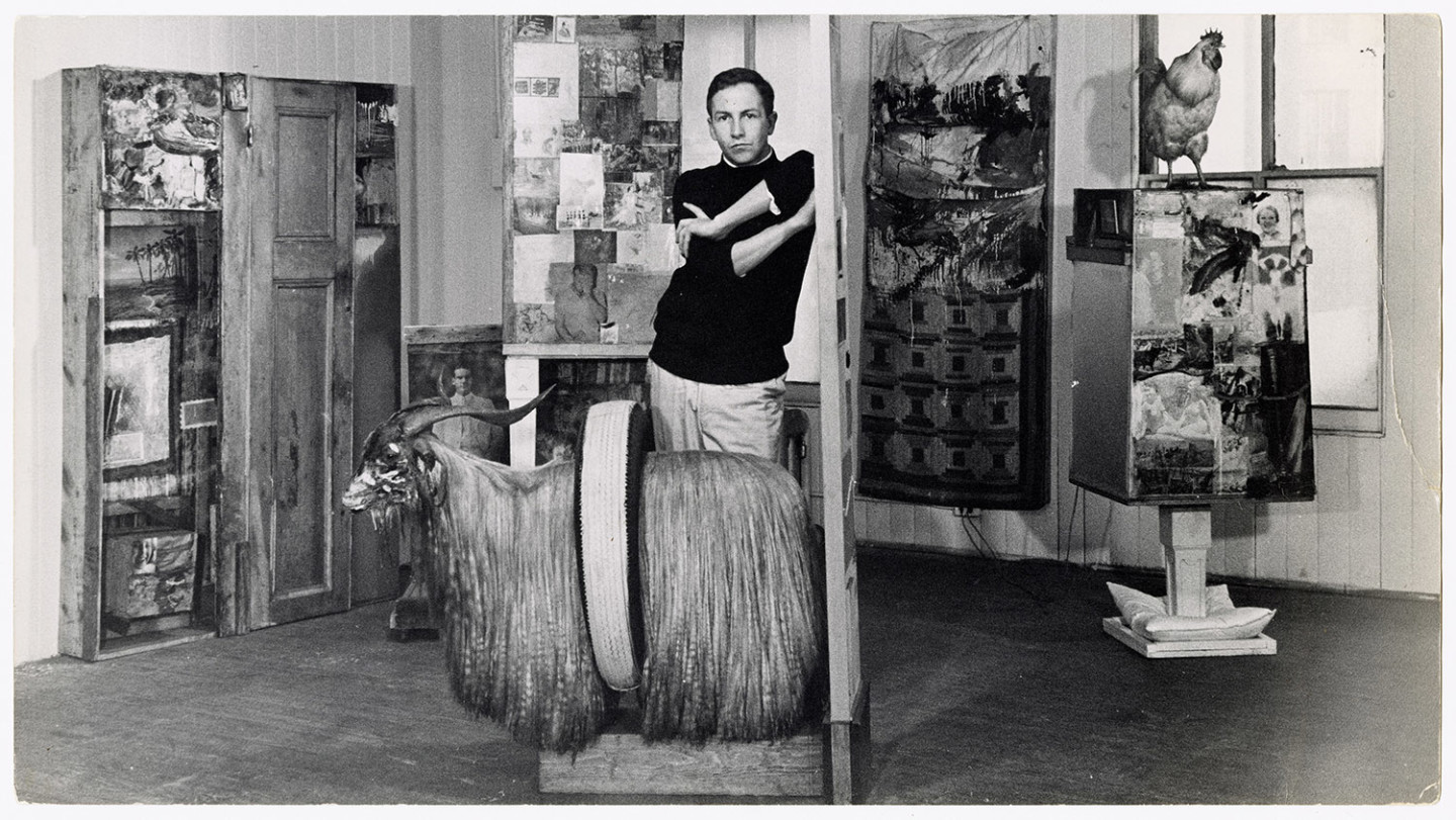 Black-and-white photo of Robert Rauschenberg standing with arms crossed behind "Monogram"